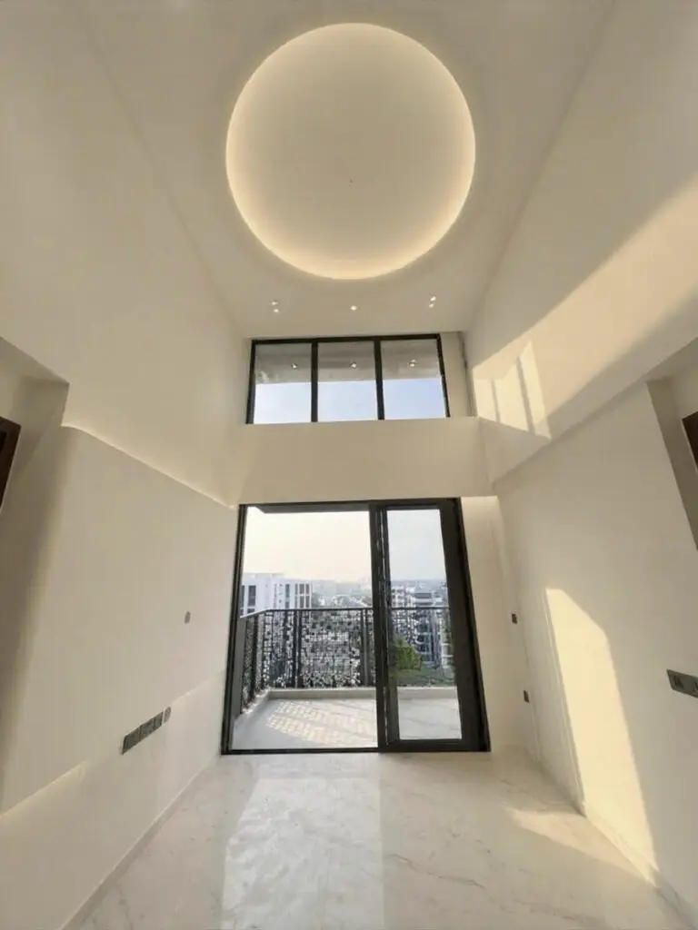 A bright, modern high-ceiling penthouse living room featuring polished white marble flooring and crisp white walls. On the left, a textured beige wall-mounted TV panel with concealed top and bottom LED backlighting features electrical outlets and a media cable port. Floor-to-ceiling white sheer curtains cover a large window or balcony glass door, above which sits a high clerestory window section. The soaring double-volume ceiling features a striking circular recessed cove light with warm illumination, small recessed spotlights, and a modern white ceiling fan suspended by a long downrod.