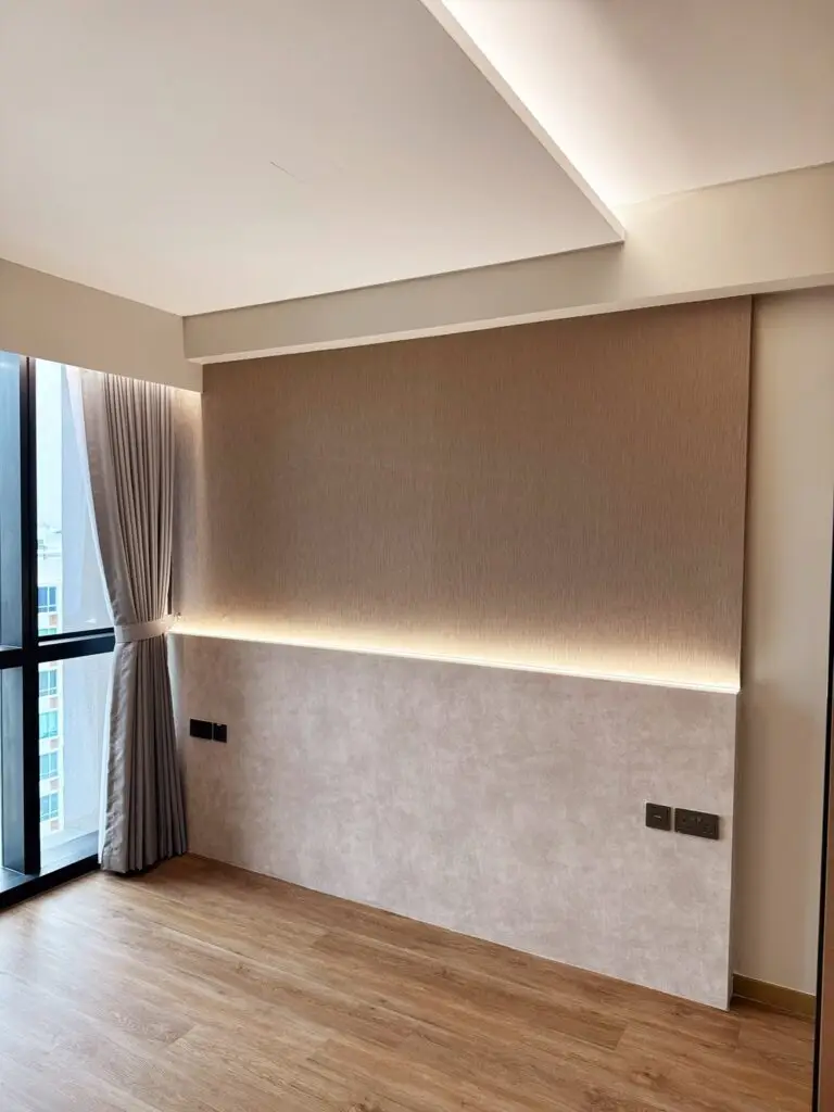 A modern master bedroom featuring warm wood-look laminate flooring, white walls, and a multi-tiered false ceiling with concealed warm perimeter cove lighting. Against the main wall, a two-tone custom bedhead panel combines a textured upper brown wood-grain panel and a lighter concrete-textured lower section, separated by a horizontal LED strip light providing soft accent glow. The bedhead panel is fitted with dark electrical sockets and switches on either side. To the left, full-height windows feature vertical grey curtains tied back neatly, and to the right, an open doorway reveals a glimpse of an interior hallway
