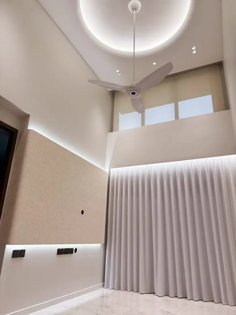 A bright, modern high-ceiling penthouse living room featuring polished white marble flooring and crisp white walls. On the left, a textured beige wall-mounted TV panel with concealed top and bottom LED backlighting features electrical outlets and a media cable port. Floor-to-ceiling white sheer curtains cover a large window or balcony glass door, above which sits a high clerestory window section. The soaring double-volume ceiling features a striking circular recessed cove light with warm illumination, small recessed spotlights, and a modern white ceiling fan suspended by a long downrod.